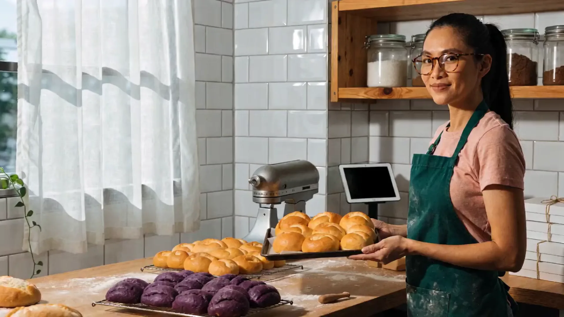 The baker in her home bakery holding a tray of fresh pandesal, smiling, with ube rolls cooling on the worktable and the tablet on its stand.