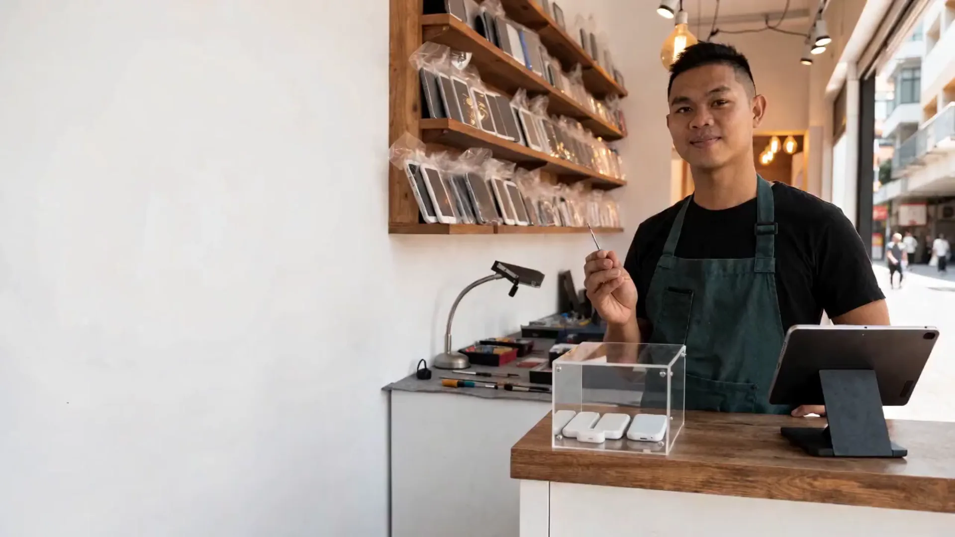 The technician behind the counter of his phone and computer repair shop, smiling, with bagged devices on the shelf and his workbench behind him.