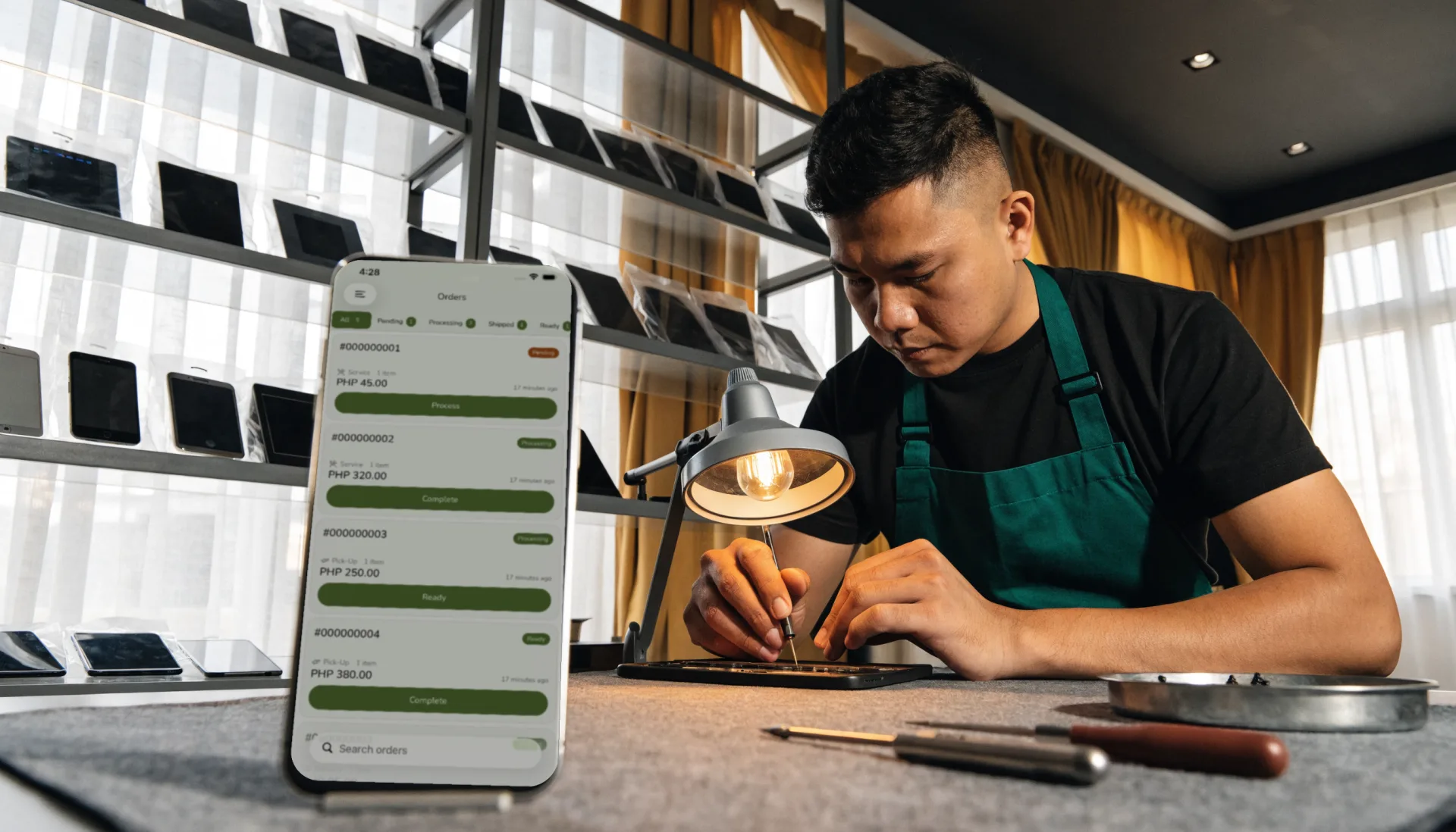 The technician works on an opened phone under the bench lamp, the job queue by status on Fuze Store on a phone standing beside him.